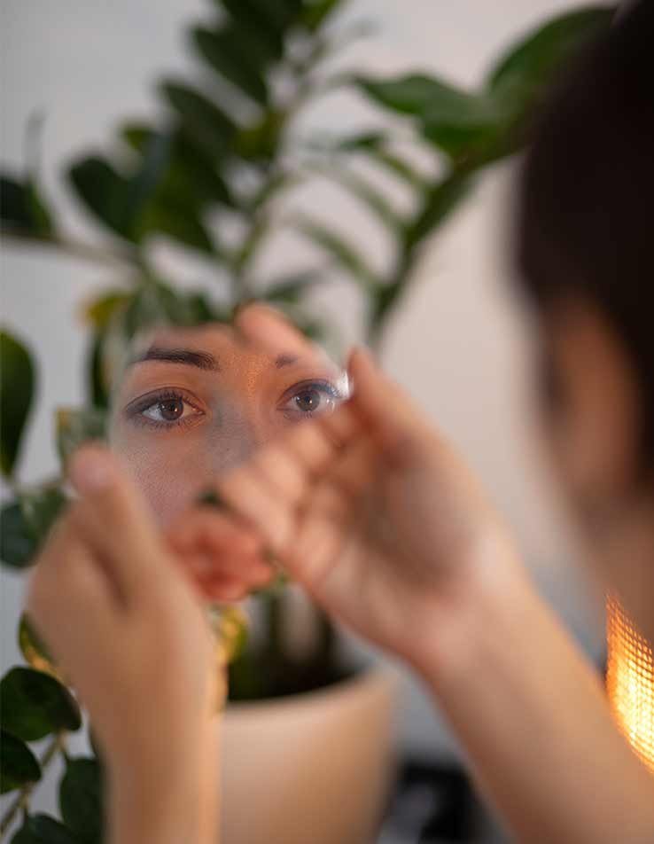 Mulher segurando espelho partido, com olhos intensos refletidos entre folhas verdes, em momento de introspecção.
