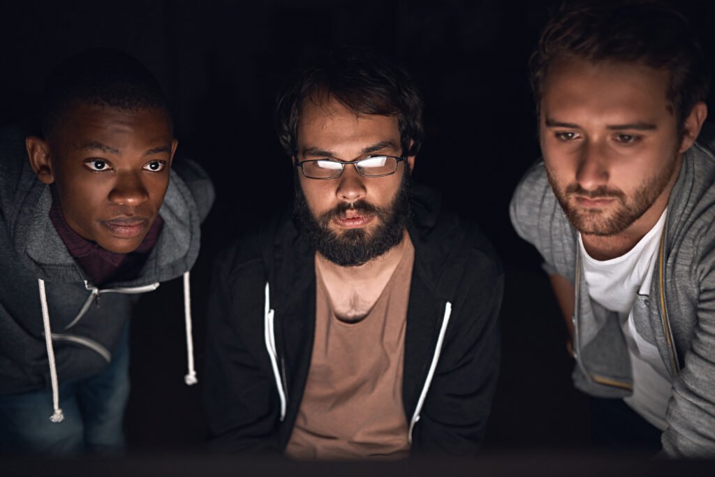 Três homens focados a olhar para um ecrã, a trabalhar em conjunto numa sala escura, simbolizando colaboração, concentração e resolução de problemas.